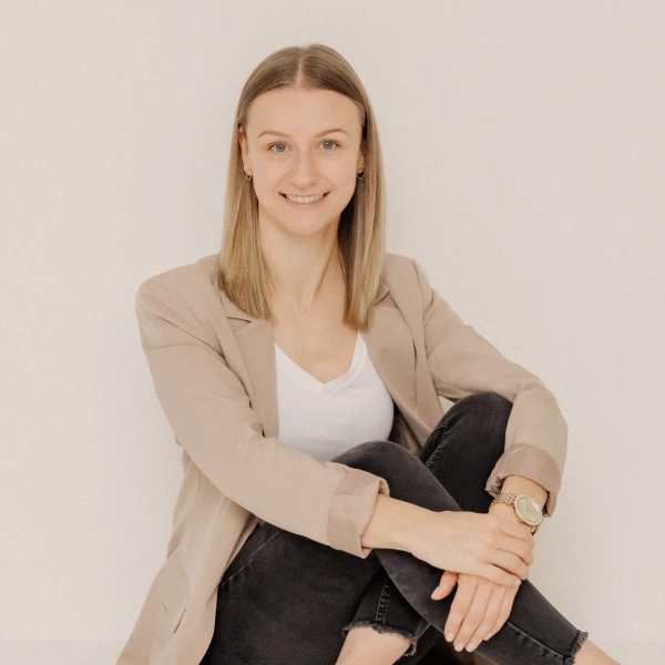 Leonie Rieder, eine junge Frau mit dunkelblonden Haaren, sitzt auf dem Boden und lächelt freundlich in die Kamera. Sie trägt ein weißes Oberteil, einen hellbraunen Blazer und eine schwarze Hose.