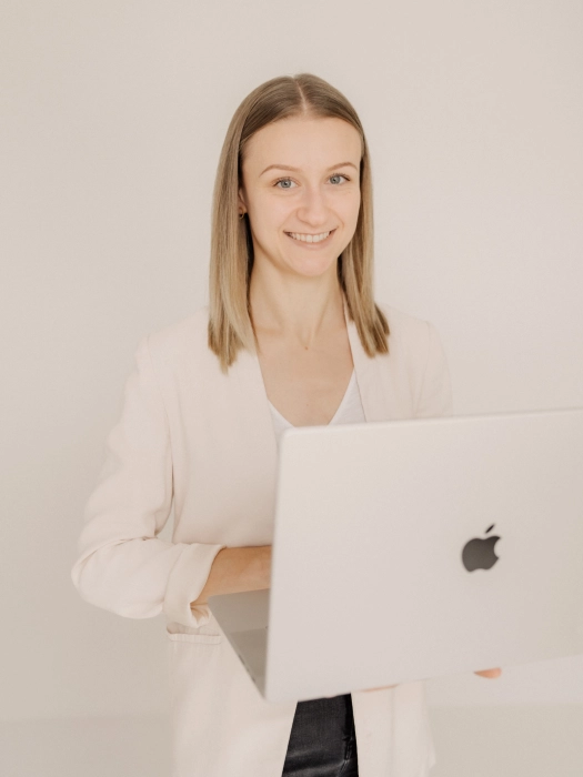 Leonie-Rieder-WordPress-Expertin-Eifel-3 Leonie Rieder, eine junge Frau mit dunkelblonden Haaren, hat ein Laptop in der Hand und lächelt freundlich in de Kamera. Sie trägt ein weißes Oberteil, einen hellbeigen Blazer und eine schwarze Hose.