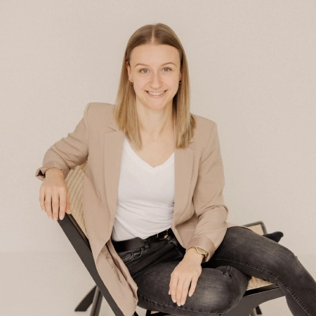 WordPress-Webdesignerin-Leonie-Rieder-2 Leonie Rieder, eine junge Frau mit dunkelblonden Haaren, hat ein Laptop in der Hand und lächelt freundlich in de Kamera. Sie trägt ein weißes Oberteil, einen hellbeigen Blazer und eine schwarze Hose.